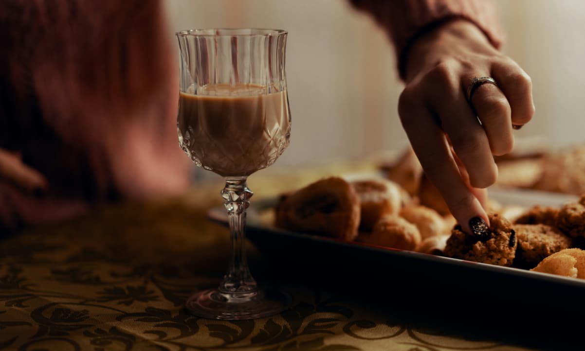 an ornate glass filled with creamy brown liquid. a hand to the right wearing a diamond ring is picking up a cookie from a plate