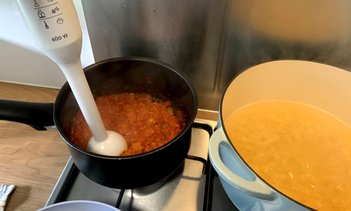 two pots: one containing pasta, and one containing a red sauce being blended with an immersion belnder