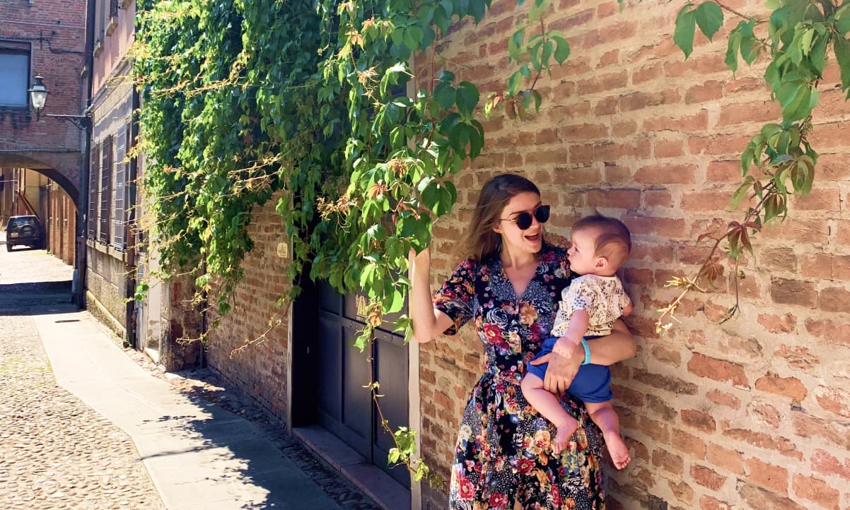 a woman in a floral dress holding a baby in a cobbled Italian street with hanging green vines