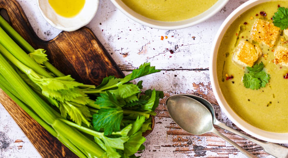 various items arranged on a white, rustic tabletop, including a wooden chopping board, fresh celery, two soup spoons, and a bowl of vegetable soup