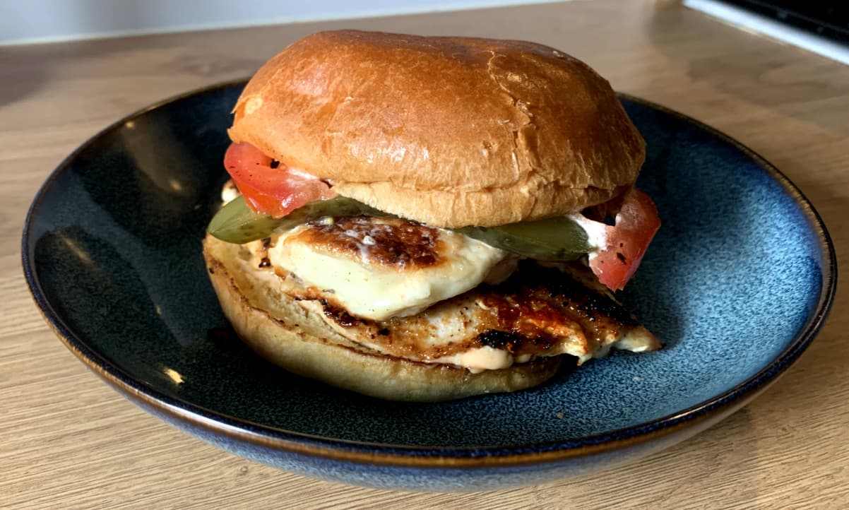 chicken and halloumi burger on a brioche bun with sliced tomato and pickle poking out the sides, on a blue plate