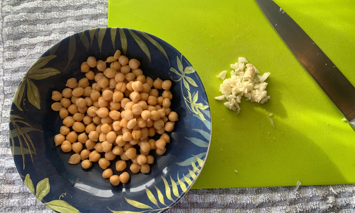 a bowl of cooked, unseasoned chickpeas in a blue bowl, and chopped garlic on a green chopping board in the background