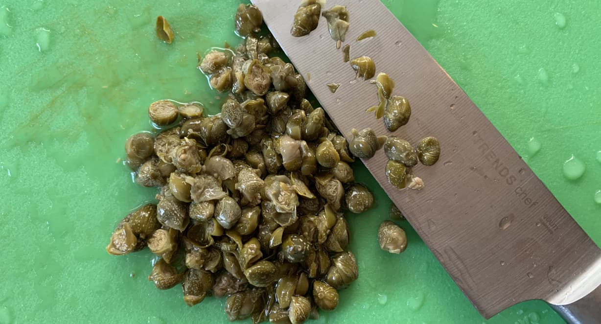 a large knife chopping capers on a green chopping board