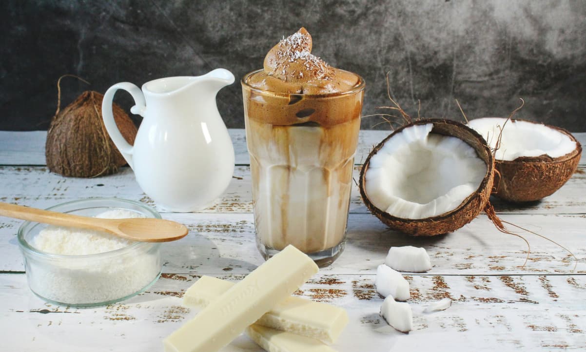 fresh coconut halves, a brown and white smoothie in a glass, and a white mulk jug atop a distressed white table