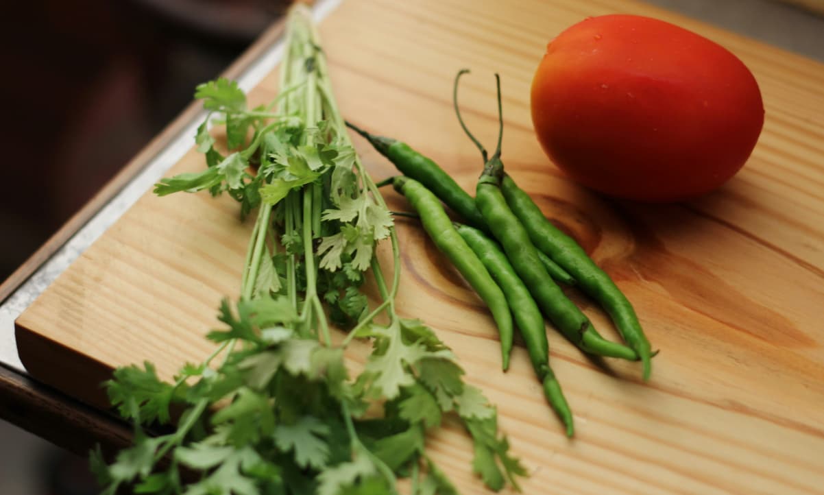 coriander stalks, fresh green chillies and a single tomato on a wooden chopping board