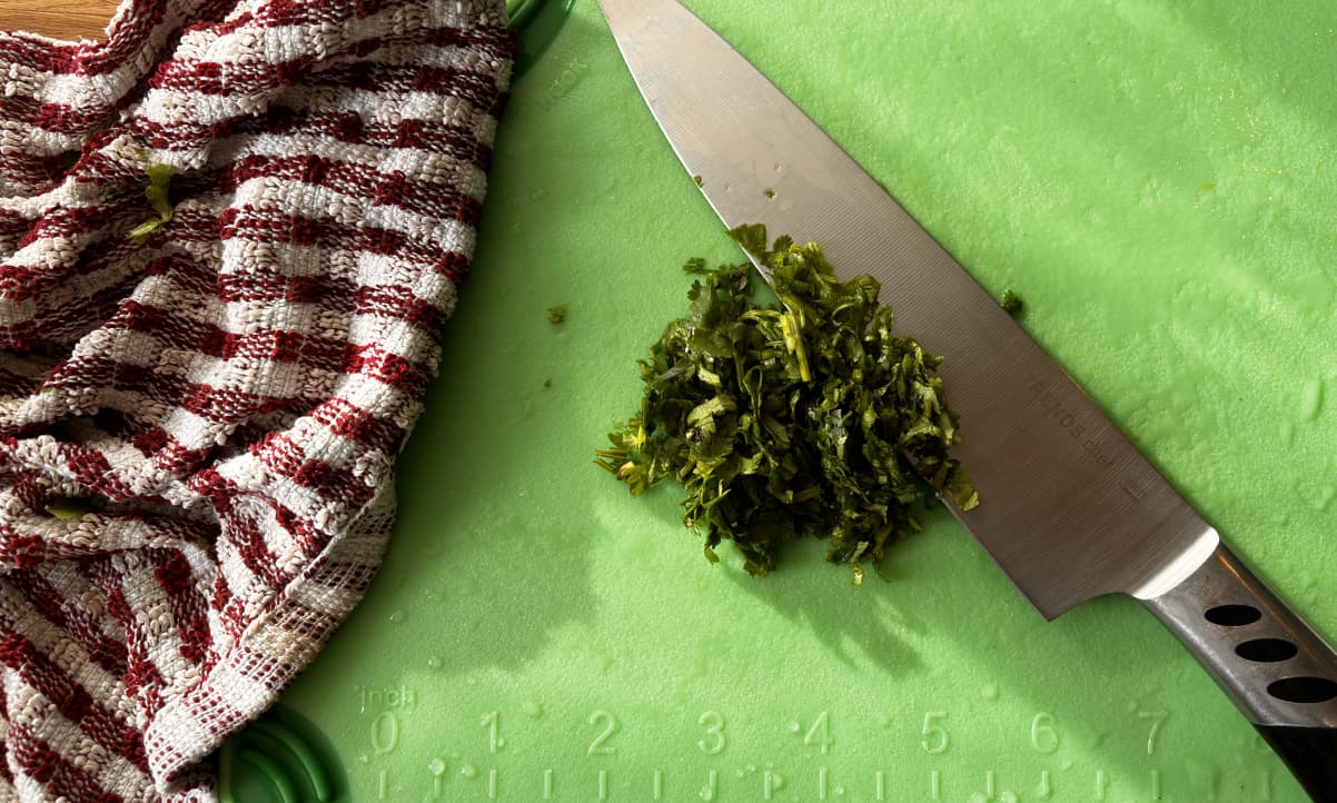 chopped coriander stems and leaves on a green chopping board, being chopped by a large knife