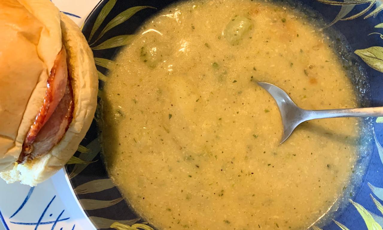 cheesy courgette and potato soup in a blue bowl decorated with green leaves, and a bacon roll on the side