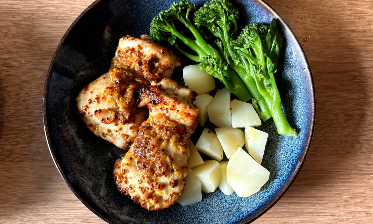 two roasted chicken thighs with wholegrain mustard sauce on top, in a blue bowl with boiled potatoes and boiled broccoli