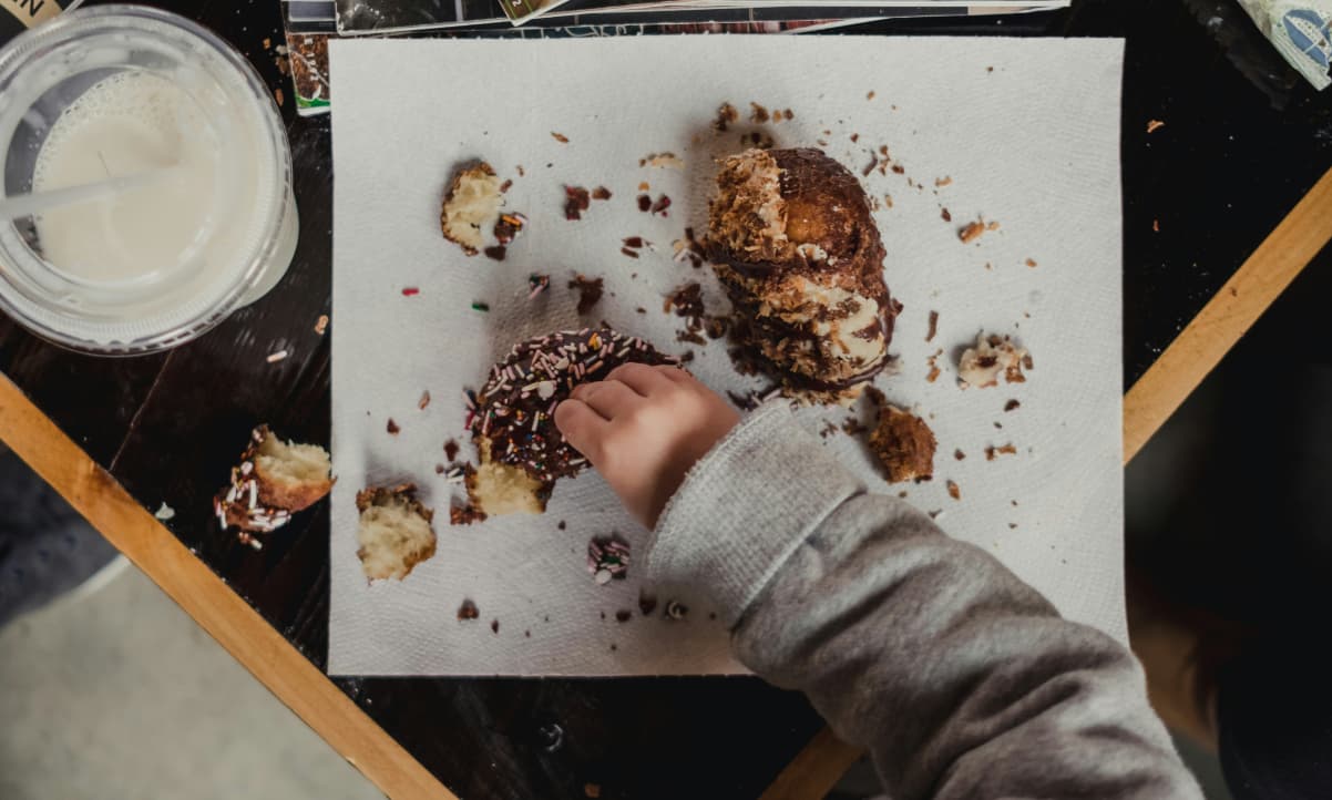 a baby's hand reaching for a doughnut on a table covered with doughnut crumbs