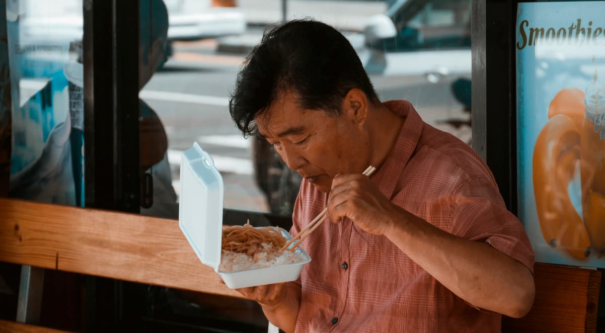 a middle aged asian man eating noodles and rice from a while polystyrene container in the street