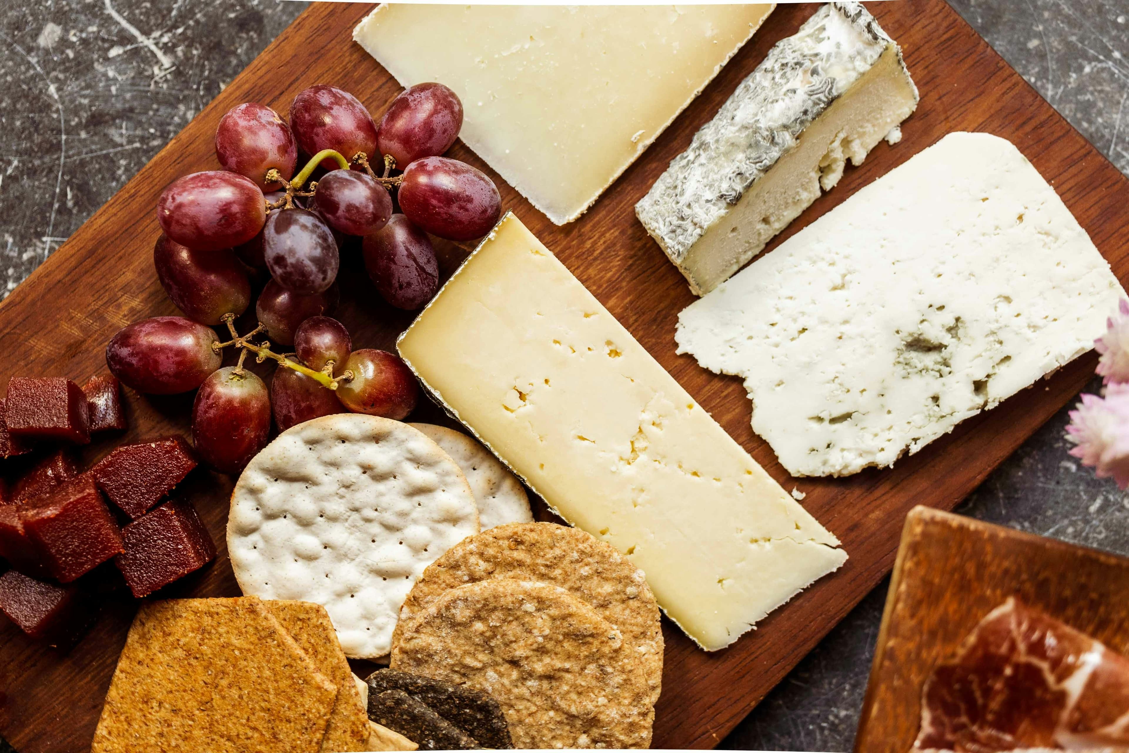 a wooden chopping board containing a variety of cheeses and crackers, and also red grapes