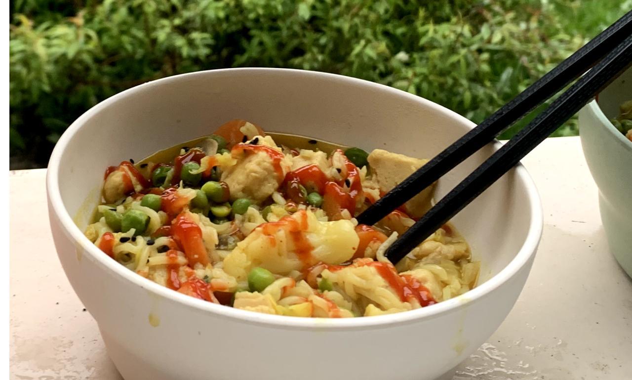 a bowl of instant ramen containing frozen vegetables and garnished with a drizzle of sriracha