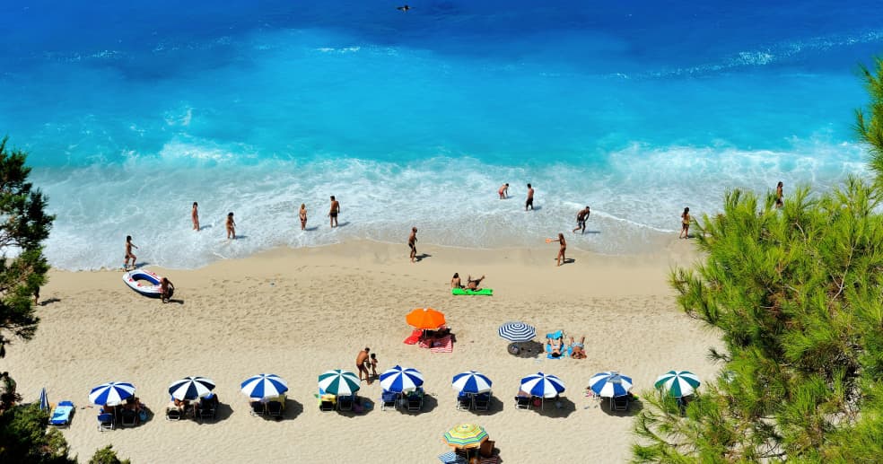 a beach photographed from above, with people in bathing suits dotted on the sand and in the shallows of the bright blue water