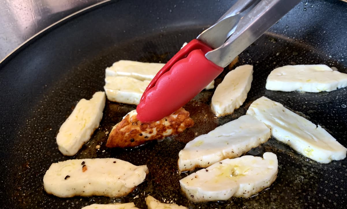 red tongs lifting the corner of a slice of halloumi frying in a pan