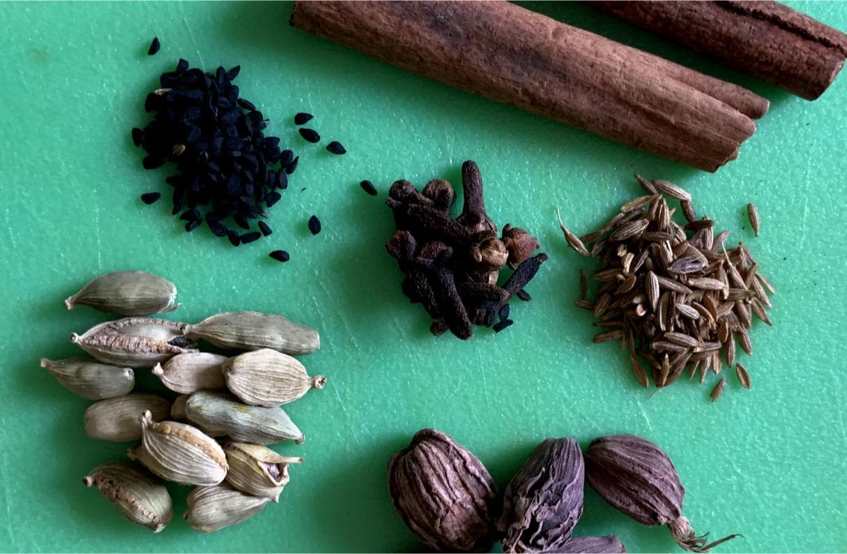 a selection of Indian whole spices arranged on a chopping board, including green cardamom, black cardamom, black onion seeds, cloves, cinnamon and cumin seeds