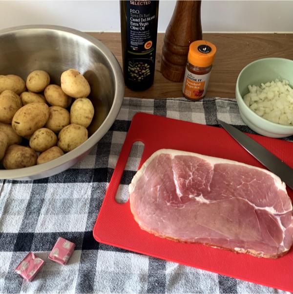 a selection of ingredients including smoked gammon, olive oil, diced onions, potatoes, paprika and stock cubes arranged on a black and white plaid cloth.