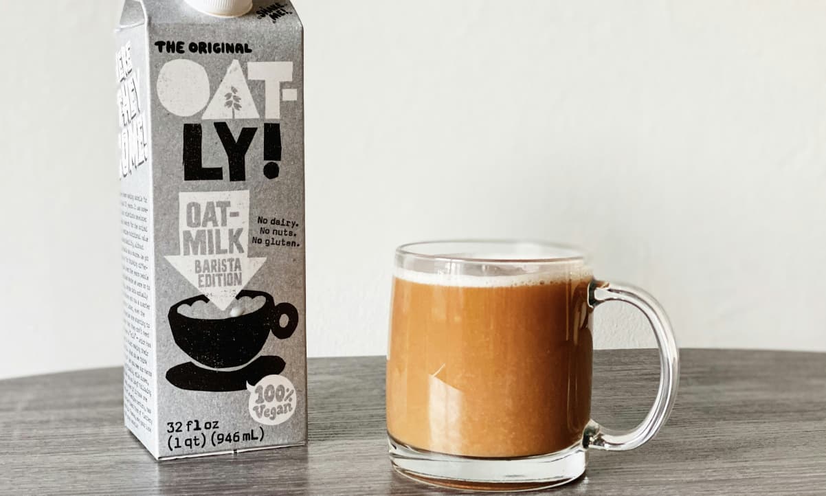 a grey carton of Oatly oat milk next to a cup of tea in a transparent mug