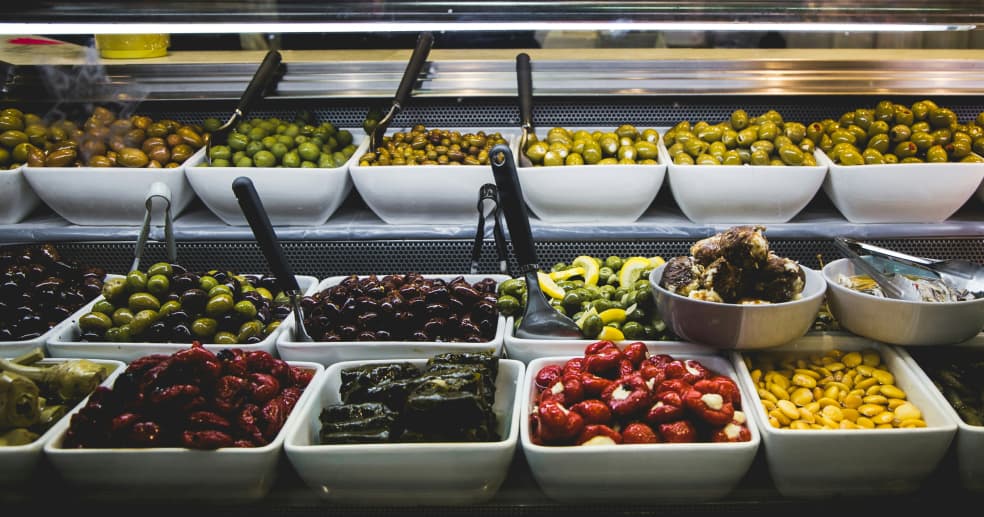 olives of various sizes and shades of green and purple at a supermarket deli counter, accompanied with other antipasto like stuffed peppers