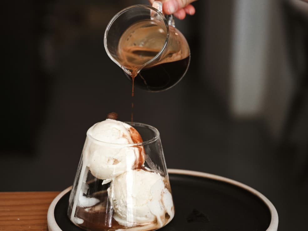 coffee being poured from a small jug onto two scoops of icecream in a glass