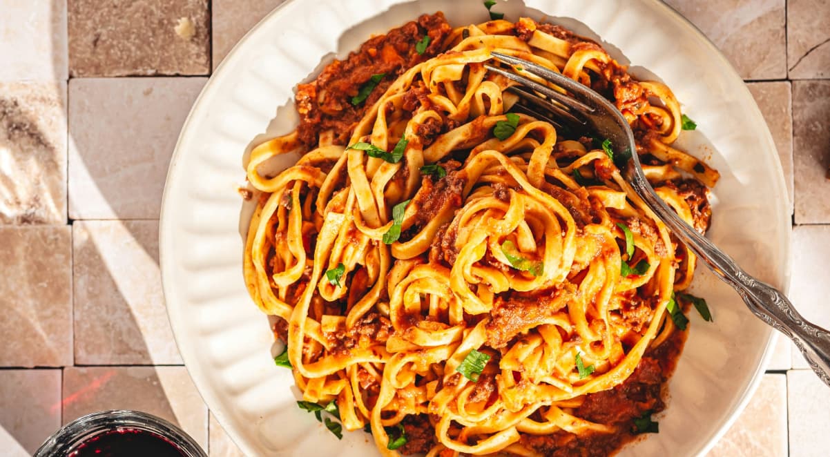 a white dinnerplate containing a large amount of pasta and meat sauce, with a fork on top