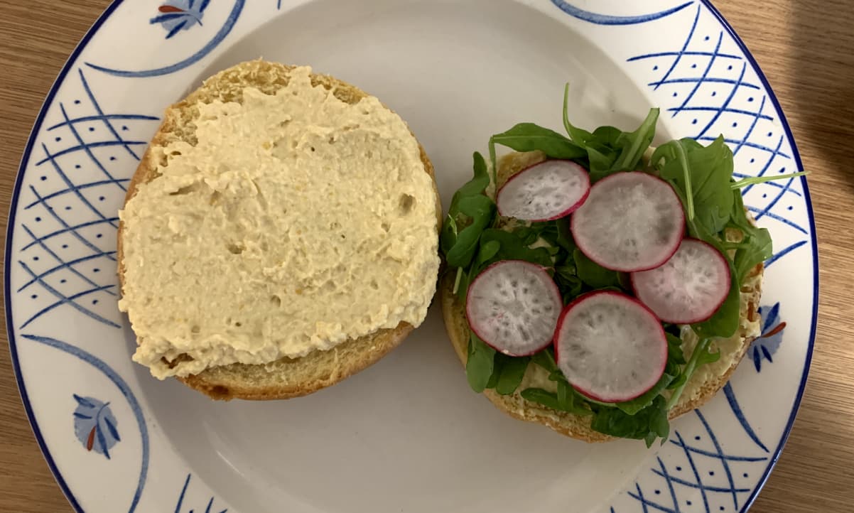 a brioche bun cut in two. one side is covered with hummus and the other with rocket and slices of radish