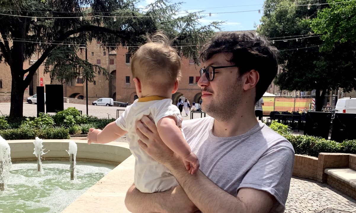 a man with dark hair and glasses holding a baby with its face turned away by a fountain in Italy