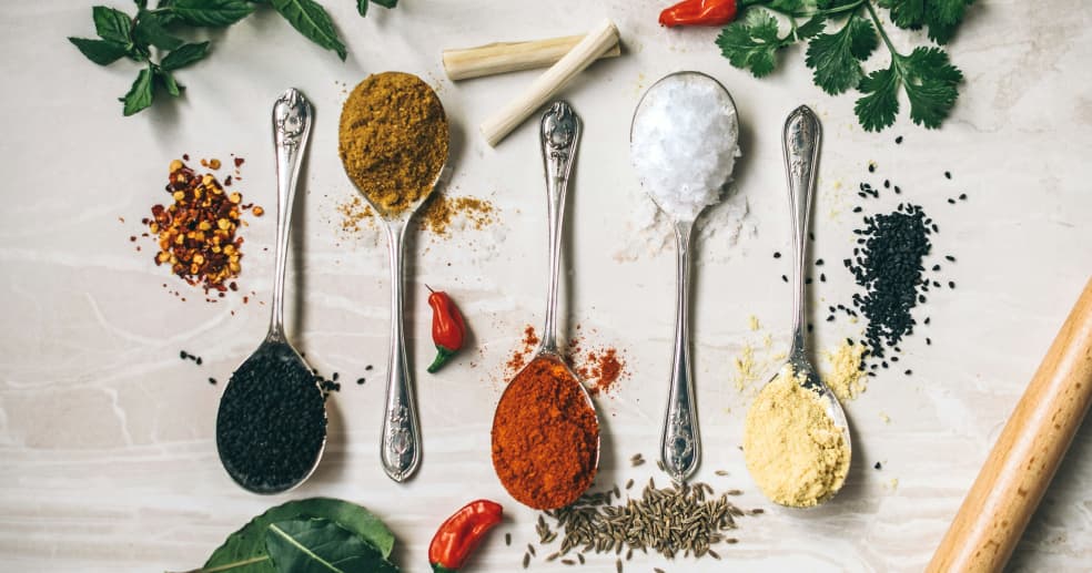 various whole and powdered spices arranged on a table, some of which are held in silver spoons