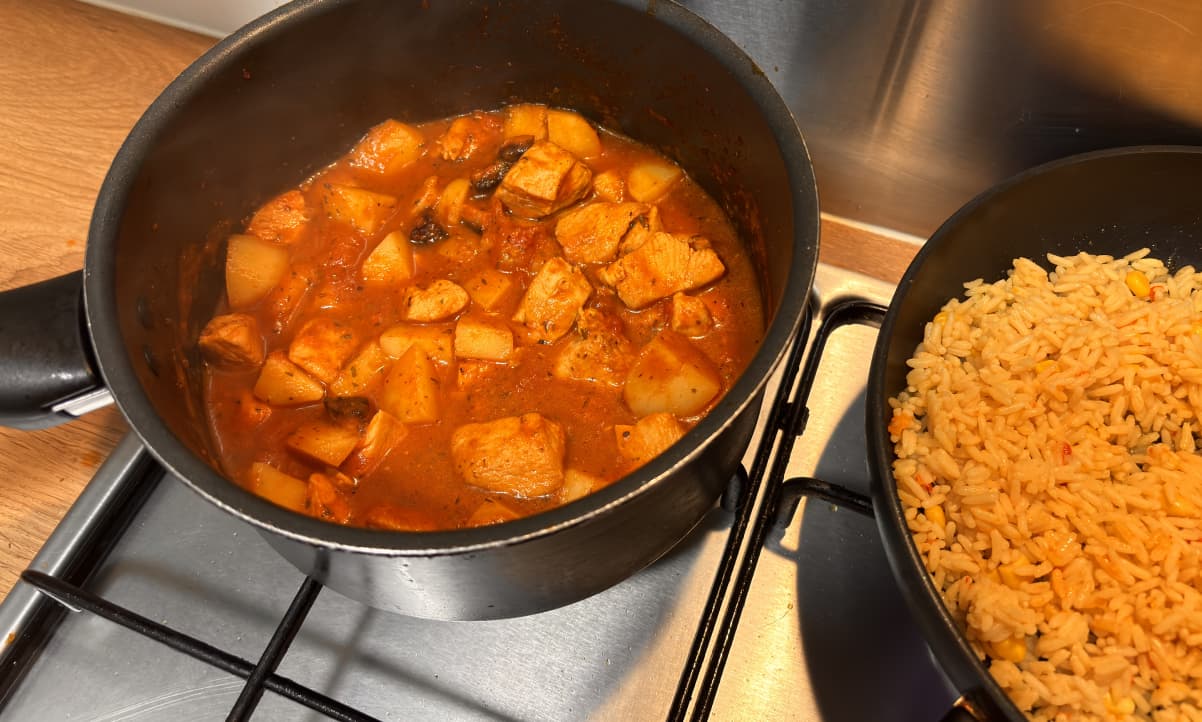 chicken and potato stew with chorizo cooking in a black pot on a gas hob, next to another pot containing yellow rice