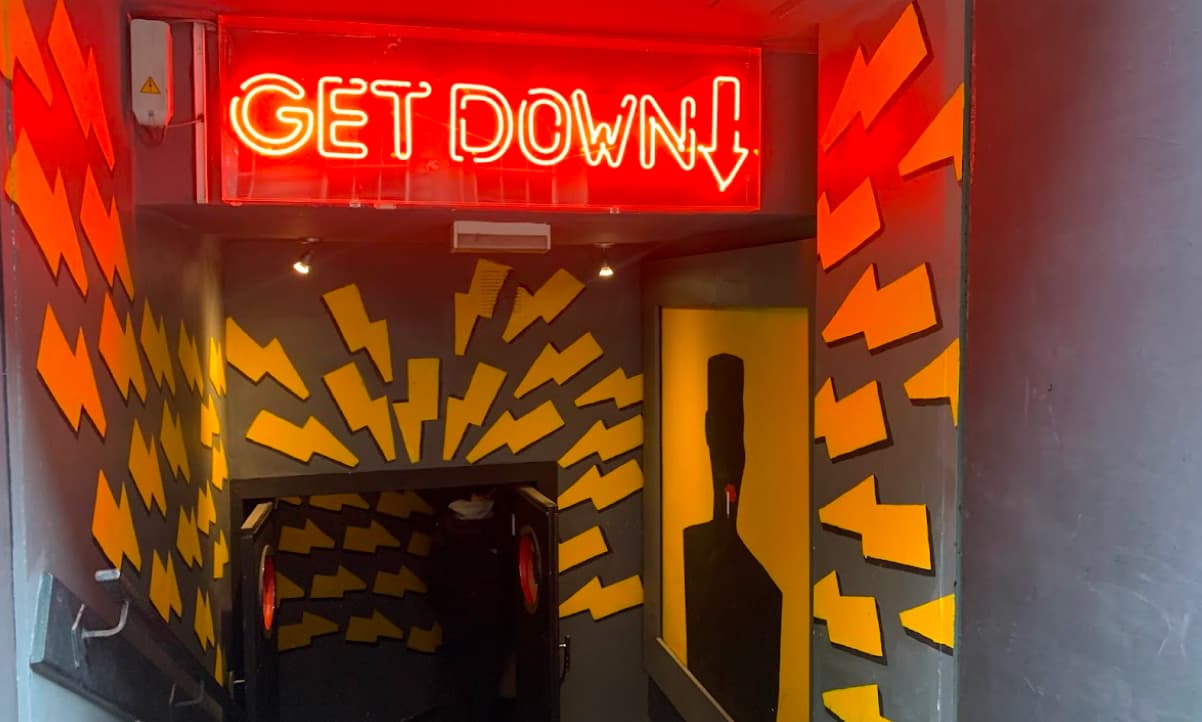 entryway to a basement venue, with a neon lit sign saying 'GET DOWN' and yellow arrow s decorating the entryway