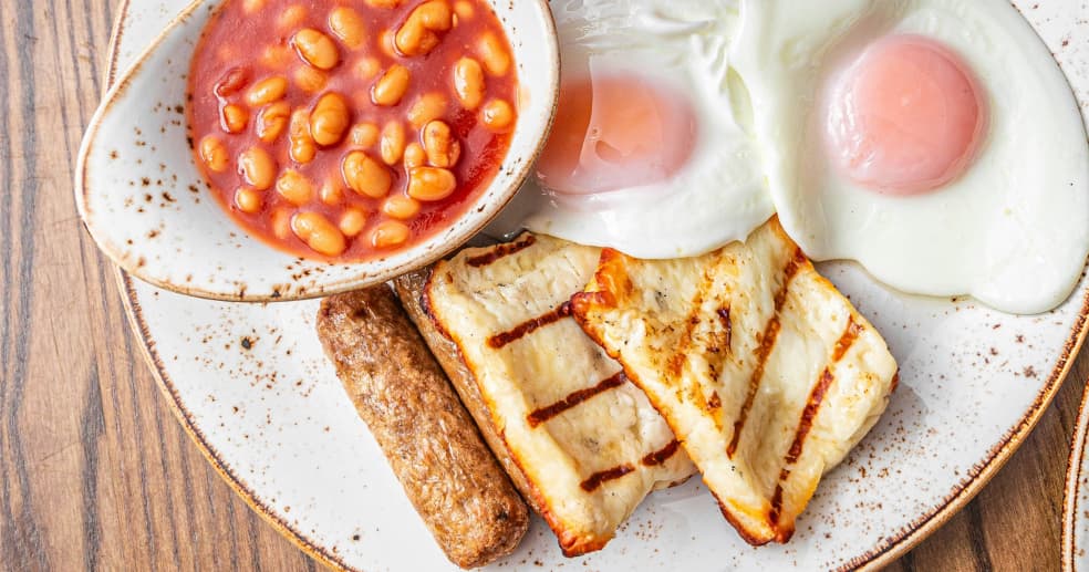 a breakfast plate filled with grilled halloumi, two fried eggs, sausages and a saucer of baked beans